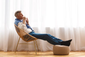 Happy senior man relaxing on chair near window, holding a cup of tea